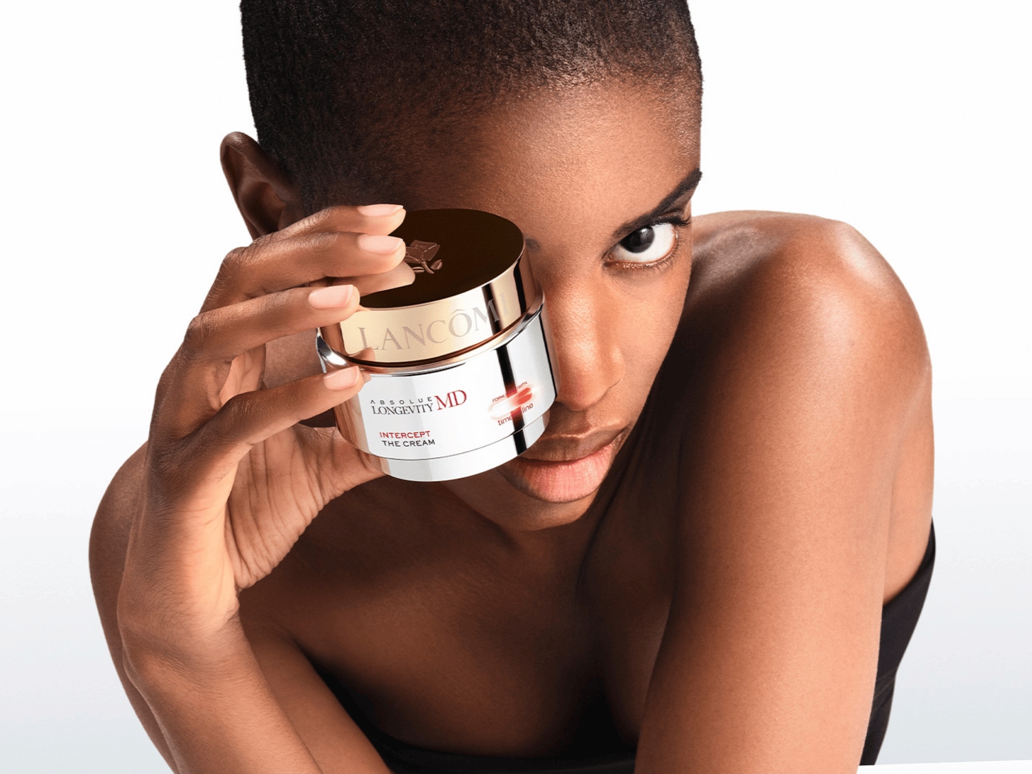 Portrét ženy držící bílý krém Lancôme Absolue Longevity MD Intercept The Cream s lesklým zlatým víčkem, který zakrývá část jejího obličeje.