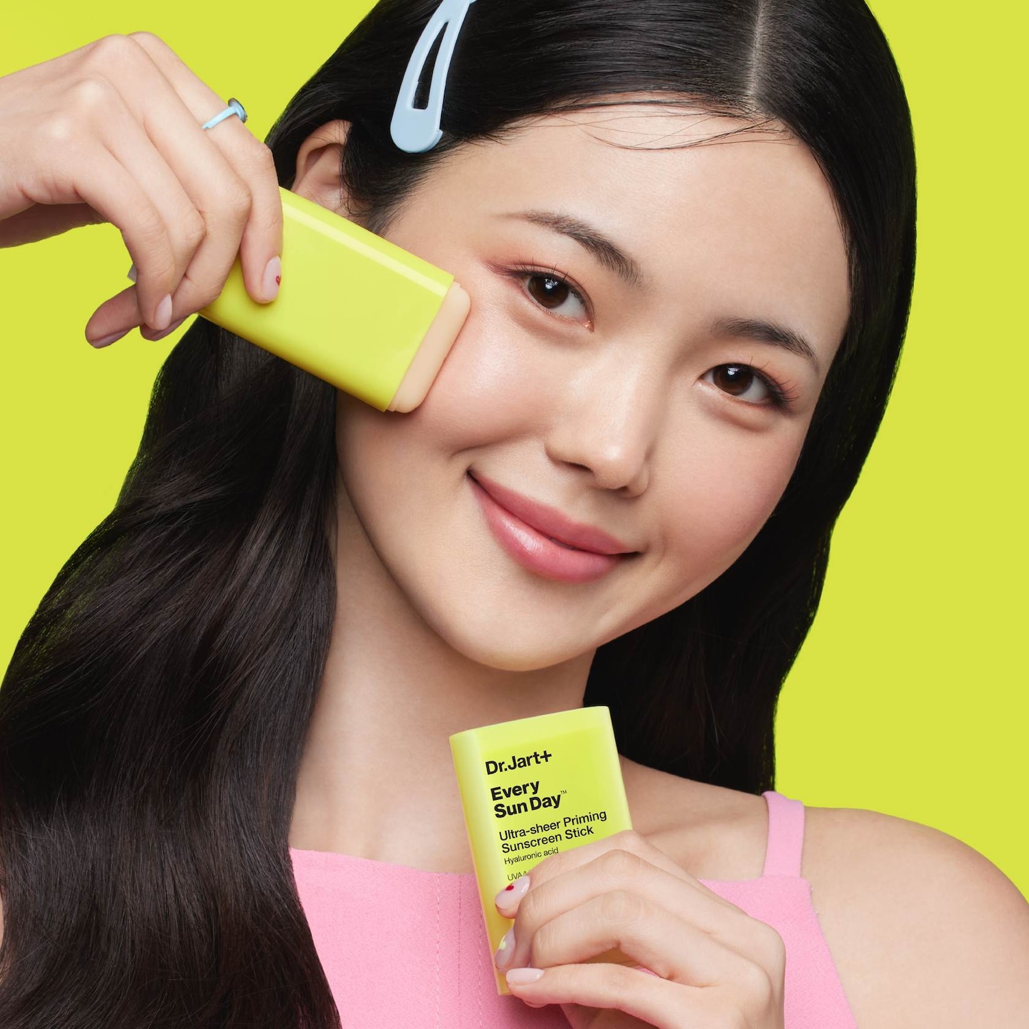 Uśmiechnięta kobieta aplikuje na twarz sztyft z filtrem przeciwsłonecznym Dr.Jart+ Every Sun Day™ Ultra-sheer Priming Sunscreen Stick z kwasem hialuronowym, zapewniający ochronę przed promieniowaniem UVA.
