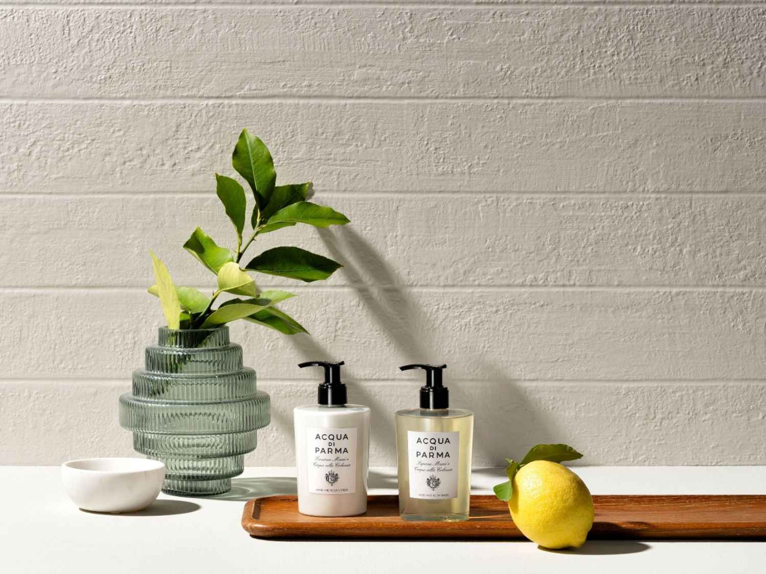 Dva produkty péče o tělo Acqua di Parma, tělové mléko a mýdlo na ruce, jsou elegantně umístěny vedle vázy se zelenými listy a citronu na dřevěném podnosu.