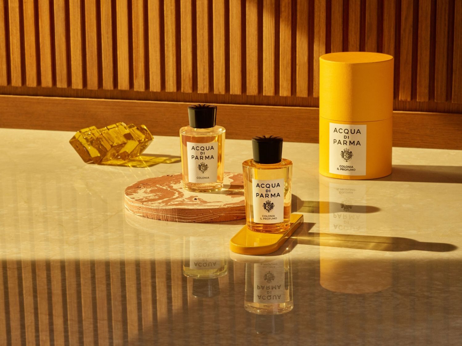 Dva flakony kolínské od Acqua di Parma stojí na mramorovém povrchu, jeden na hnědé platformě a druhý na žluté, vedle nich je žlutá krabička a žlutá skleněná dekorace.