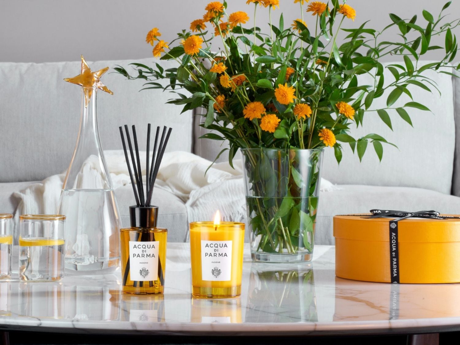 Na mramorovém stolku stojí difuzér Acqua di Parma s černými tyčinkami, žlutá svíčka, váza s oranžovými květy a dárková krabička, vše doplňuje skleněná karafa ve tvaru hvězdy.