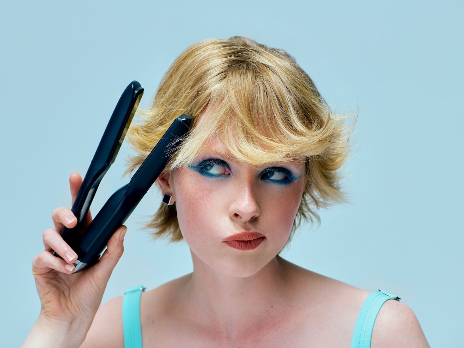 Eine Frau mit blonden, gewellten Haaren und blauem Lidschatten hält ein schwarzes Glätteisen in der Hand, um ihre Frisur zu stylen.