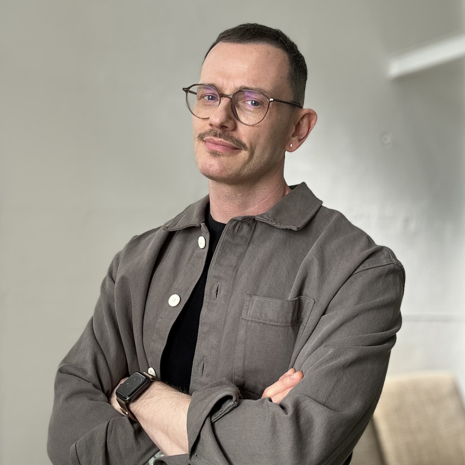 Ein Mann mit Brille, Schnurrbart und grauem Hemd steht mit verschränkten Armen da.