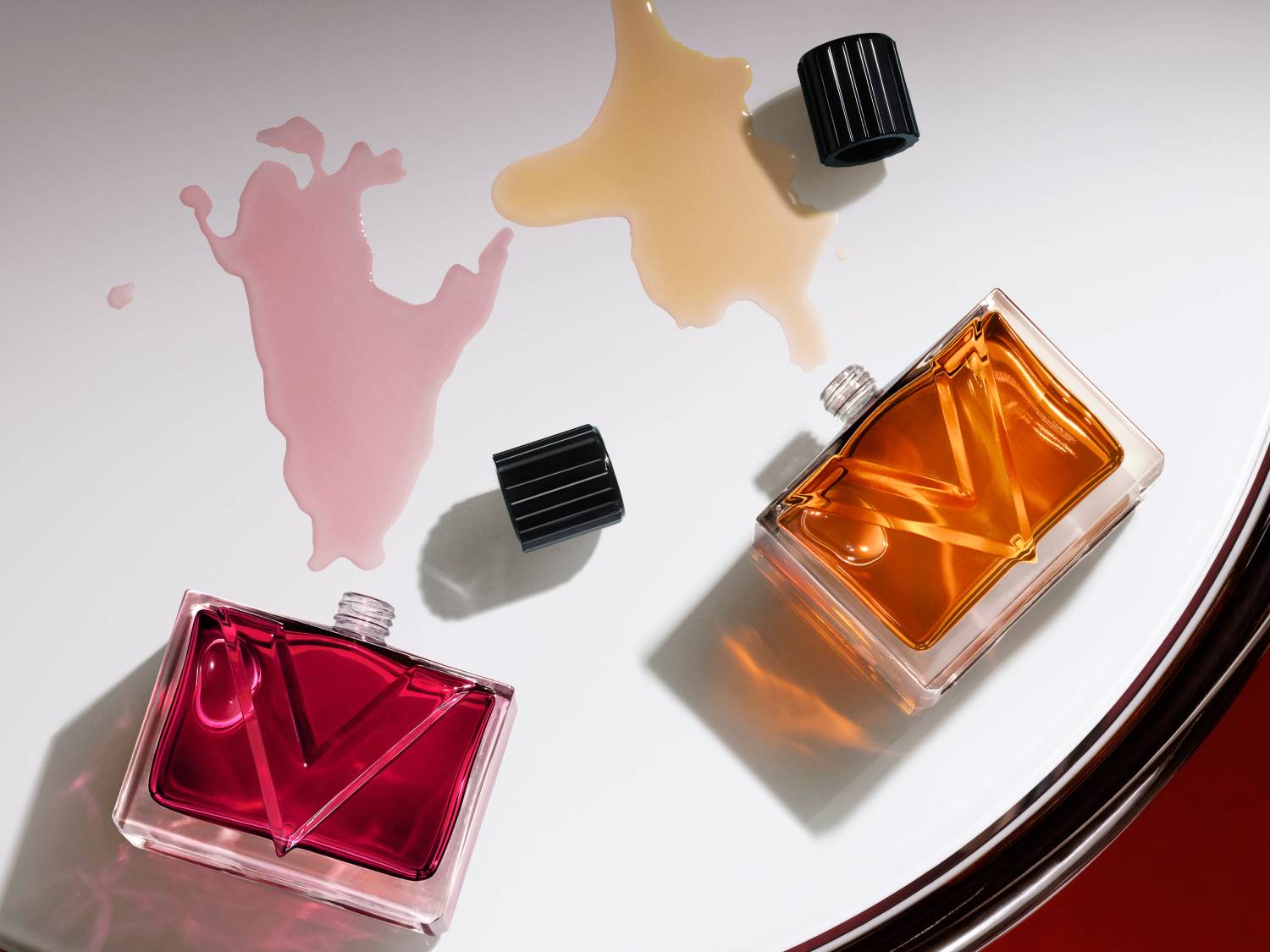 Deux flacons de parfum rectangulaires, l'un rouge et l'autre ambré, sont posés sur une surface blanche avec des éclaboussures de liquide rose et beige, et leurs capuchons noirs sont à côté.