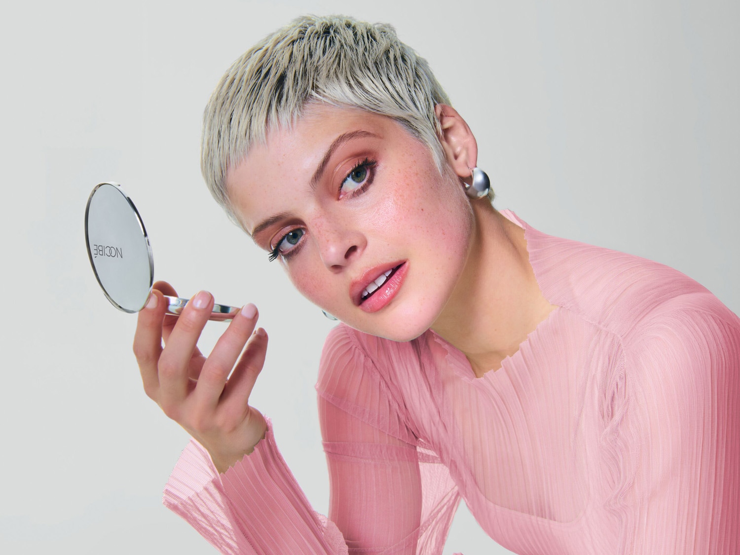 Une femme aux cheveux courts et blond platine, vêtue d'un chemisier rose, tient un poudrier Nocibé et vérifie son maquillage.