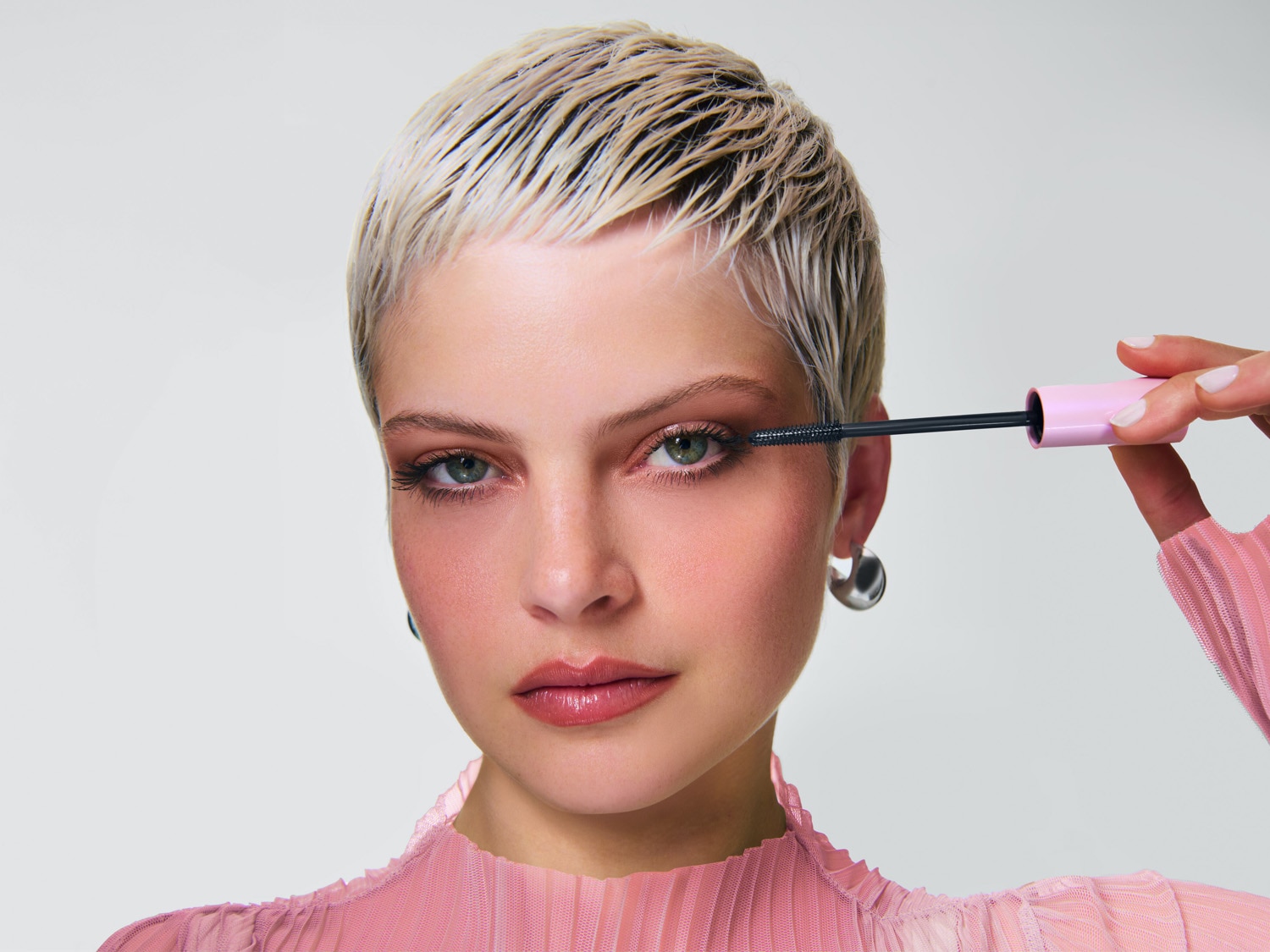 Une femme aux cheveux courts et blonds applique du mascara noir avec un applicateur rose clair, mettant en valeur ses yeux verts et son teint rosé.