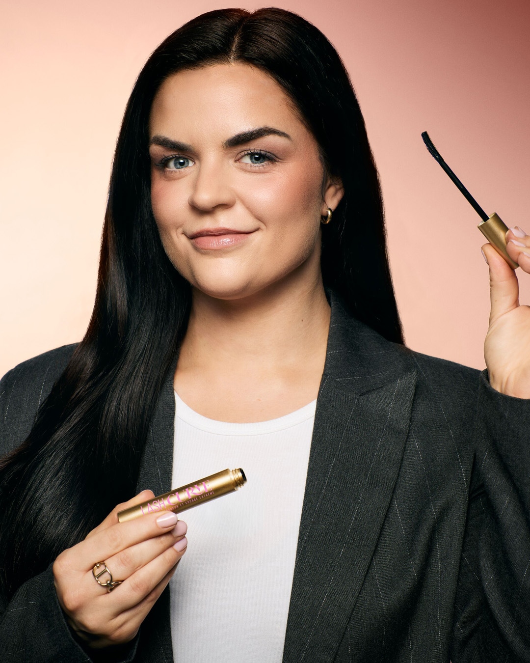 Une femme souriante tient un mascara doré Lash Curvé, le regard pétillant et les cils prêts à être sublimés.