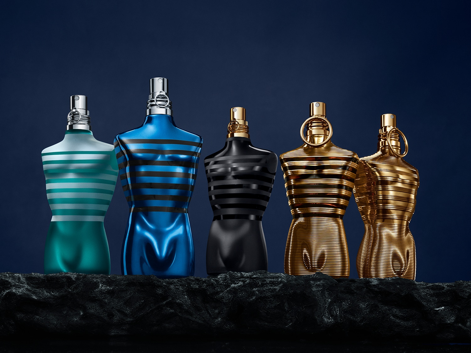 Fünf Jean Paul Gaultier Parfümflakons in Form eines männlichen Oberkörpers, die von links nach rechts in den Farben Türkis, Blau, Schwarz und Gold auf einem dunklen, strukturierten Felsen vor einem marineblauen Hintergrund angeordnet sind.