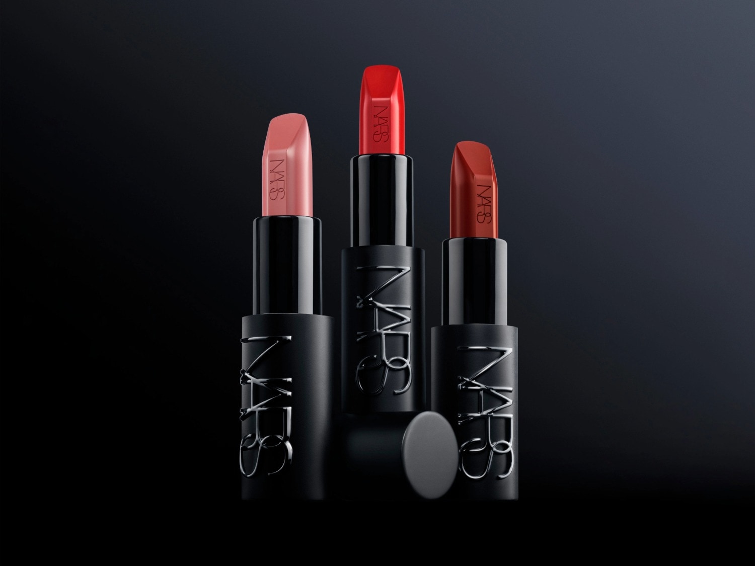 Trois rouges à lèvres NARS aux teintes rose, rouge et brique se dressent élégamment sur un fond noir, leurs étuis portant fièrement le logo argenté de la marque.