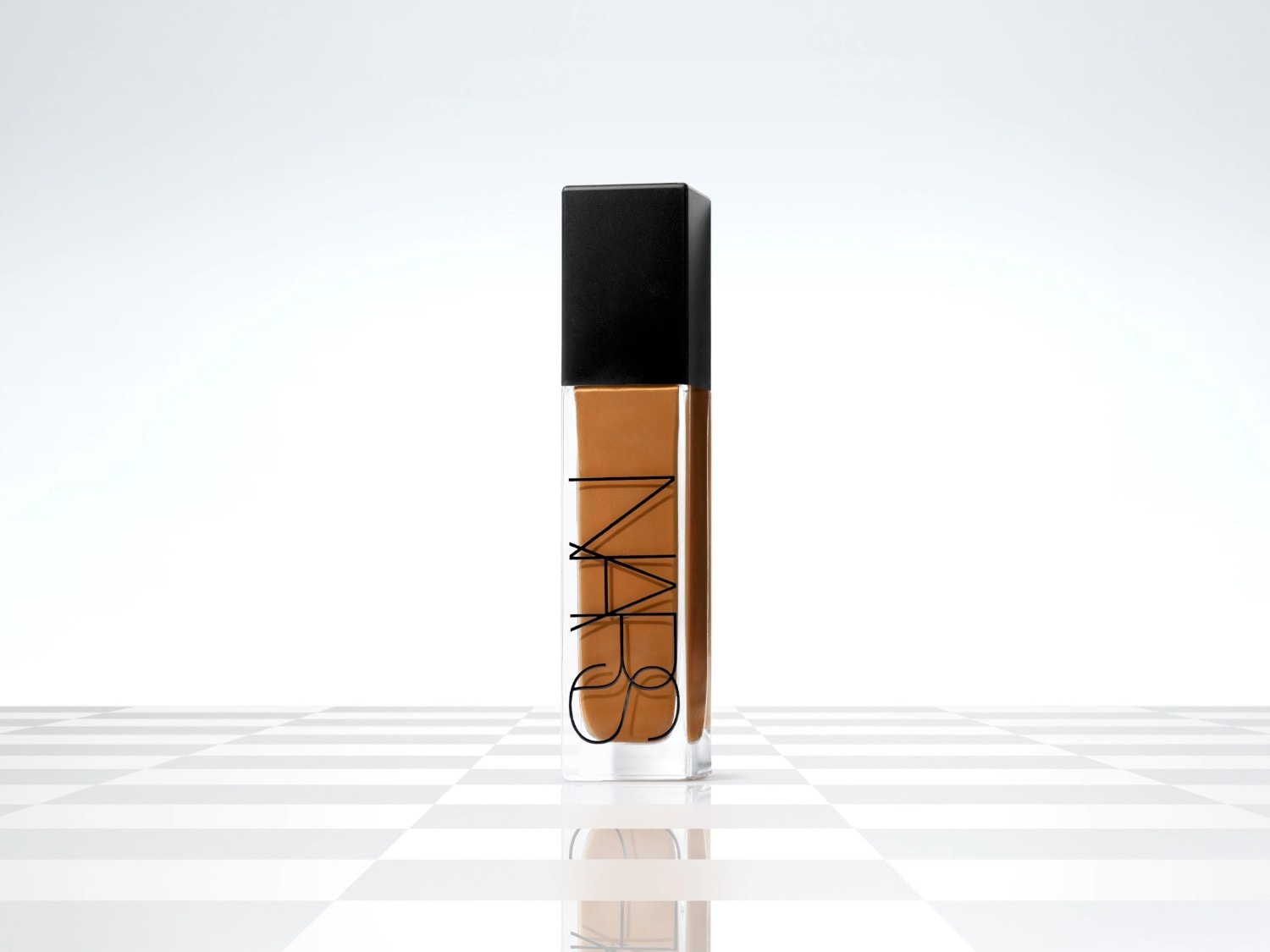 Flacon de fond de teint NARS transparent rempli de liquide marron, avec un bouchon noir, sur un fond à carreaux blancs.