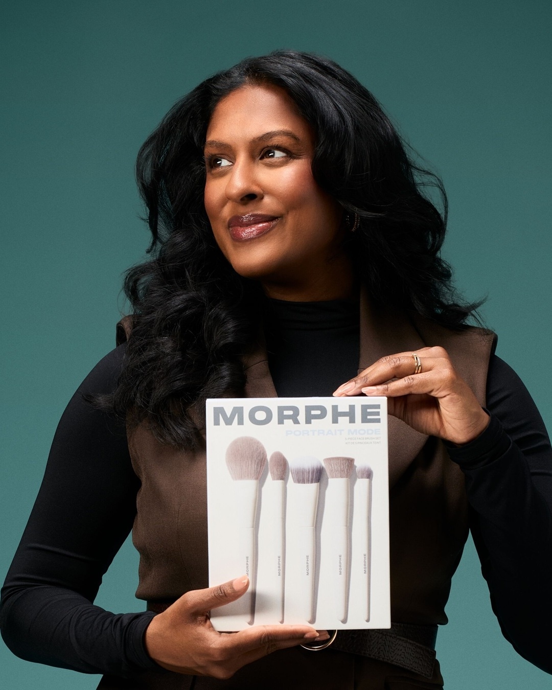 Eine Frau mit dunklen, lockigen Haaren und einem braunen Oberteil hält ein weißes Morphe Portrait Mode Pinselset in den Händen und blickt lächelnd zur Seite.