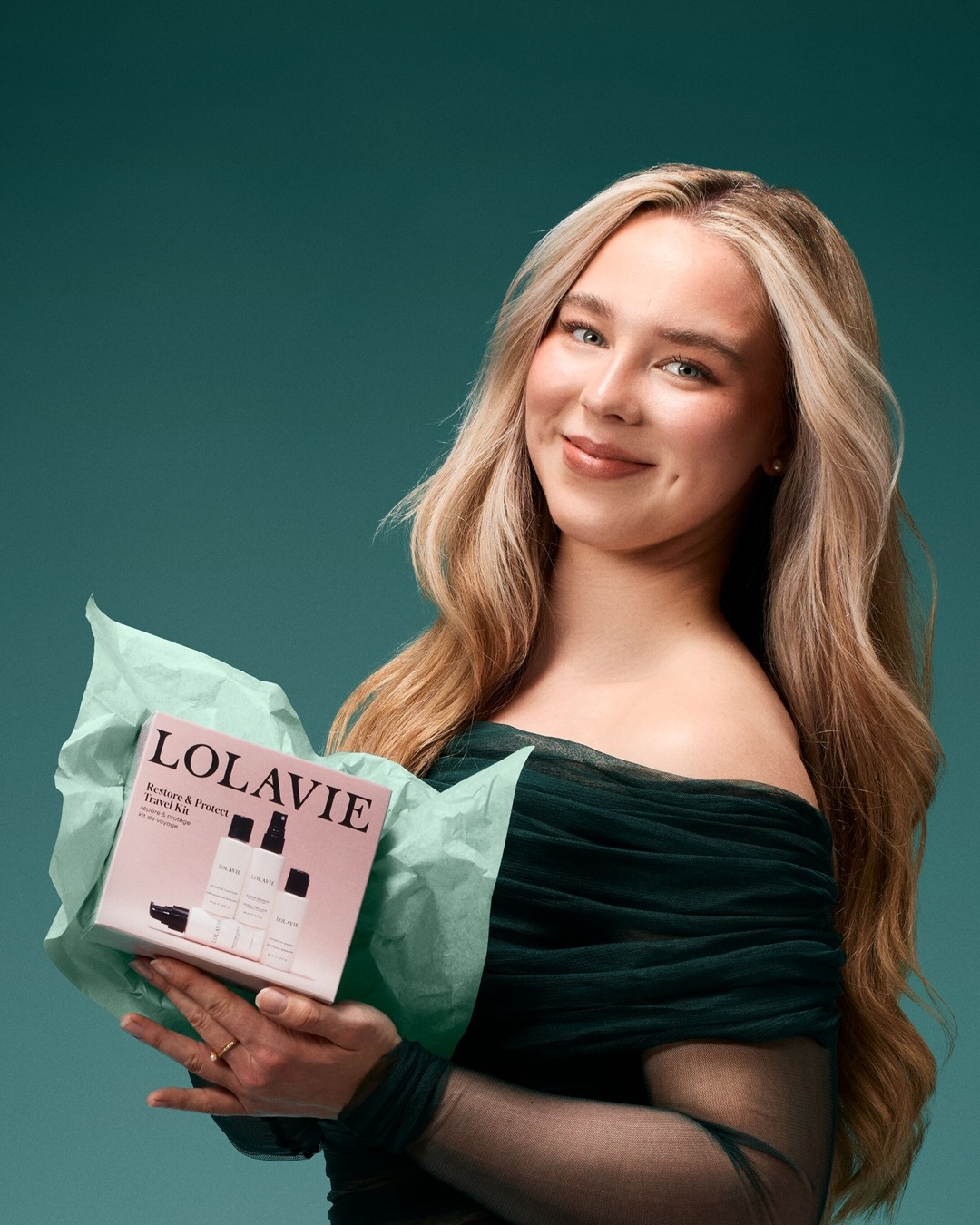 Eine lächelnde Frau mit blonden Haaren hält ein rosafarbenes Lolavie Restore & Protect Travel Kit in ihren Händen, das in mintgrünes Seidenpapier eingewickelt ist.