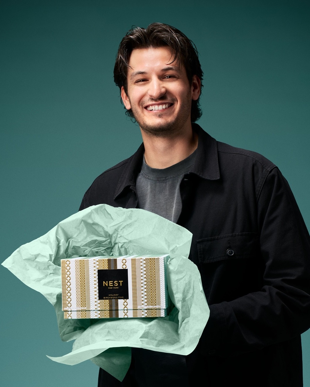Ein lächelnder Mann mit dunklem Haar und einem schwarzen Hemd hält ein Geschenk in den Händen, das in mintgrünes Seidenpapier und eine gold-weiß gemusterte Schachtel mit der Aufschrift "NEST NEW YORK HOLIDAY BIRCHWOOD PINE" eingewickelt ist.