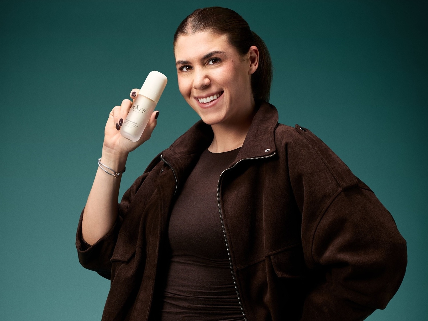 Eine lächelnde Frau mit brauner Jacke hält eine weiße Flasche des RÂYE Gentle Cream Produkts in ihrer rechten Hand.