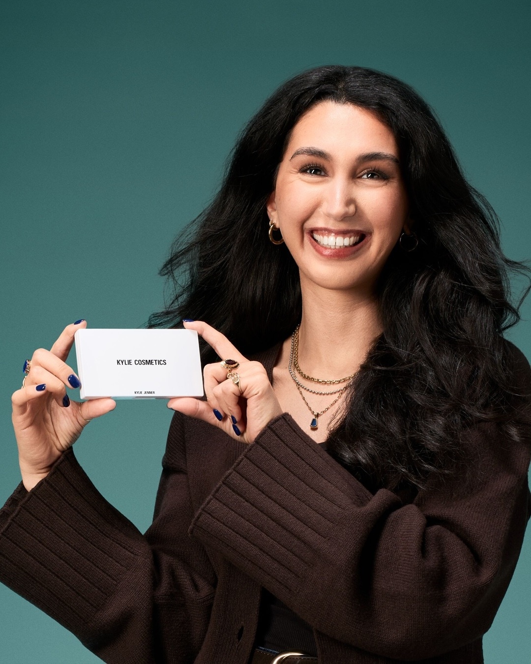 Eine lächelnde Frau mit langen, dunklen Haaren und einem braunen Strickpullover hält eine weiße Schachtel mit der Aufschrift "KYLIE COSMETICS" in ihren Händen, ihre Nägel sind dunkelblau lackiert.