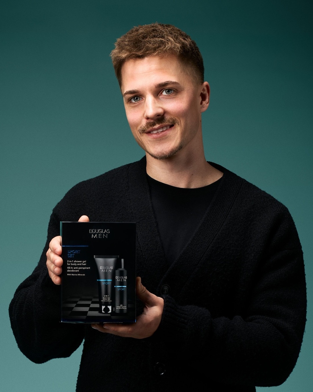 Vyras šypsosi laikydamas „Douglas Men Sport Set“ rinkinį, kuriame yra 2-in-1 dušo želė kūnui ir plaukams bei 48 valandų poveikio antiperspirantas su jūros mineralais.