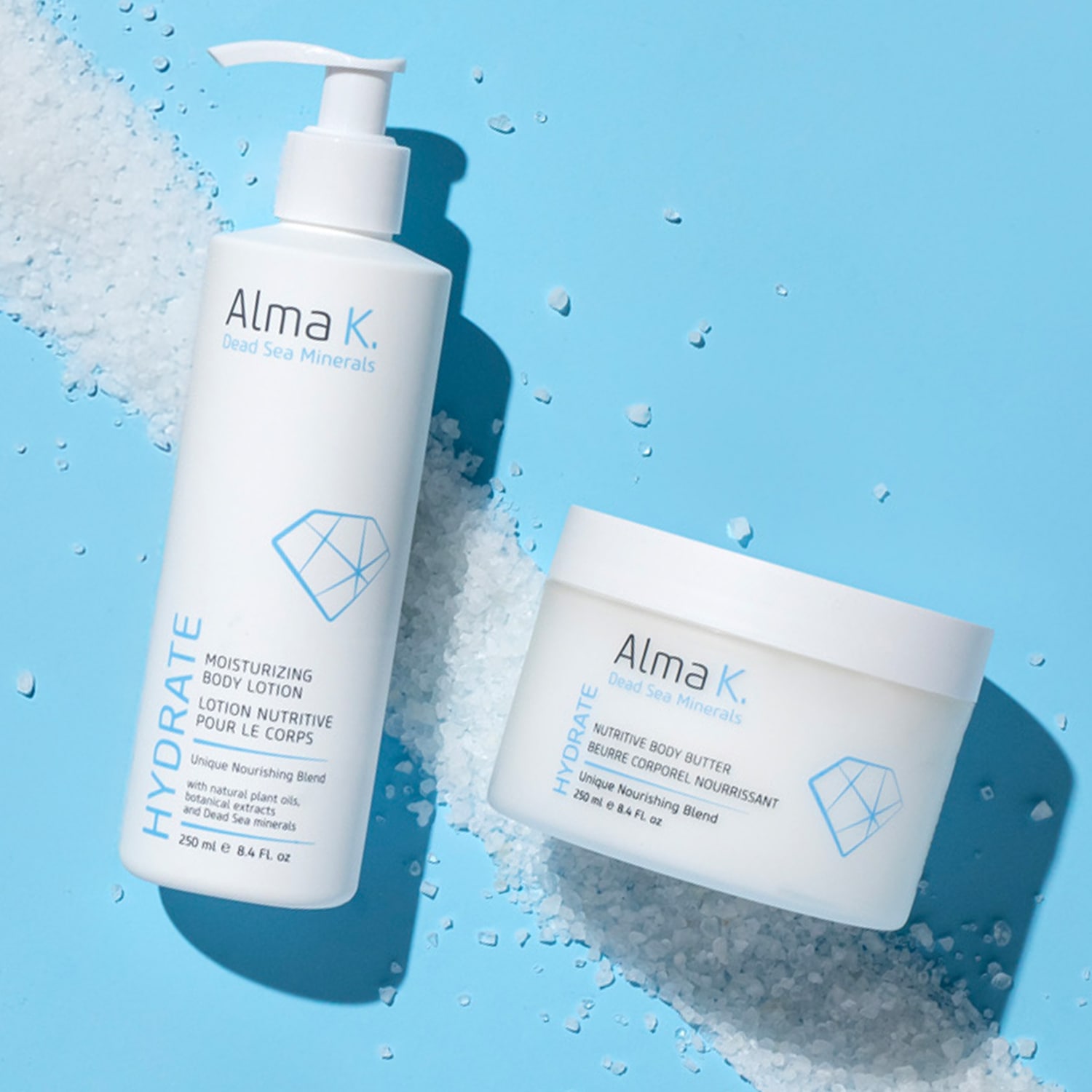 Ecco la lozione idratante per il corpo e il burro nutriente per il corpo Alma K. Dead Sea Minerals, entrambi in confezioni bianche minimaliste con accenti blu, su uno sfondo azzurro cosparso di sali minerali.