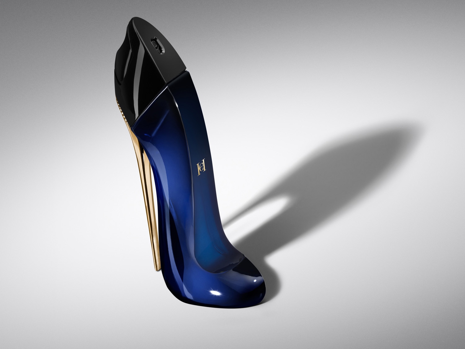 Parfumul Good Girl de la Carolina Herrera, într-o sticlă elegantă în formă de pantof cu toc înalt, de culoare albastru închis, cu un toc auriu strălucitor și logo-ul auriu "CH" pe lateral, este așezat pe o suprafață albă, cu o umbră alungită în spate.