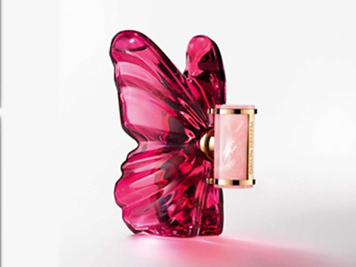 Un flacon de parfum roz, în formă de fluture, cu un design elegant și o etichetă aurie, emană o strălucire delicată pe o suprafață albă.