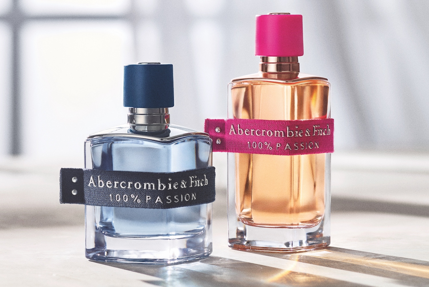 Două sticle de parfum Abercrombie & Fitch 100% Passion, una cu lichid albastru deschis și capac albastru închis, cealaltă cu lichid auriu și capac roz intens, ambele având o bandă textilă cu numele brandului și textul "100% PASSION".