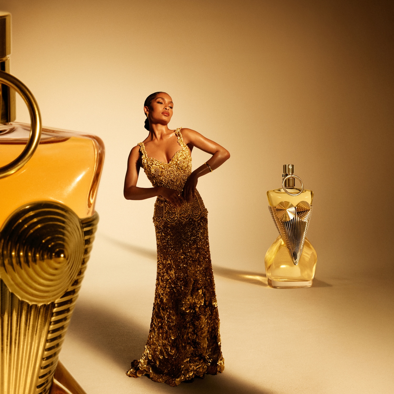 O femeie într-o rochie aurie strălucitoare pozează între două sticle de parfum Jean Paul Gaultier, ambele prezentând nuanțe aurii și designuri unice.