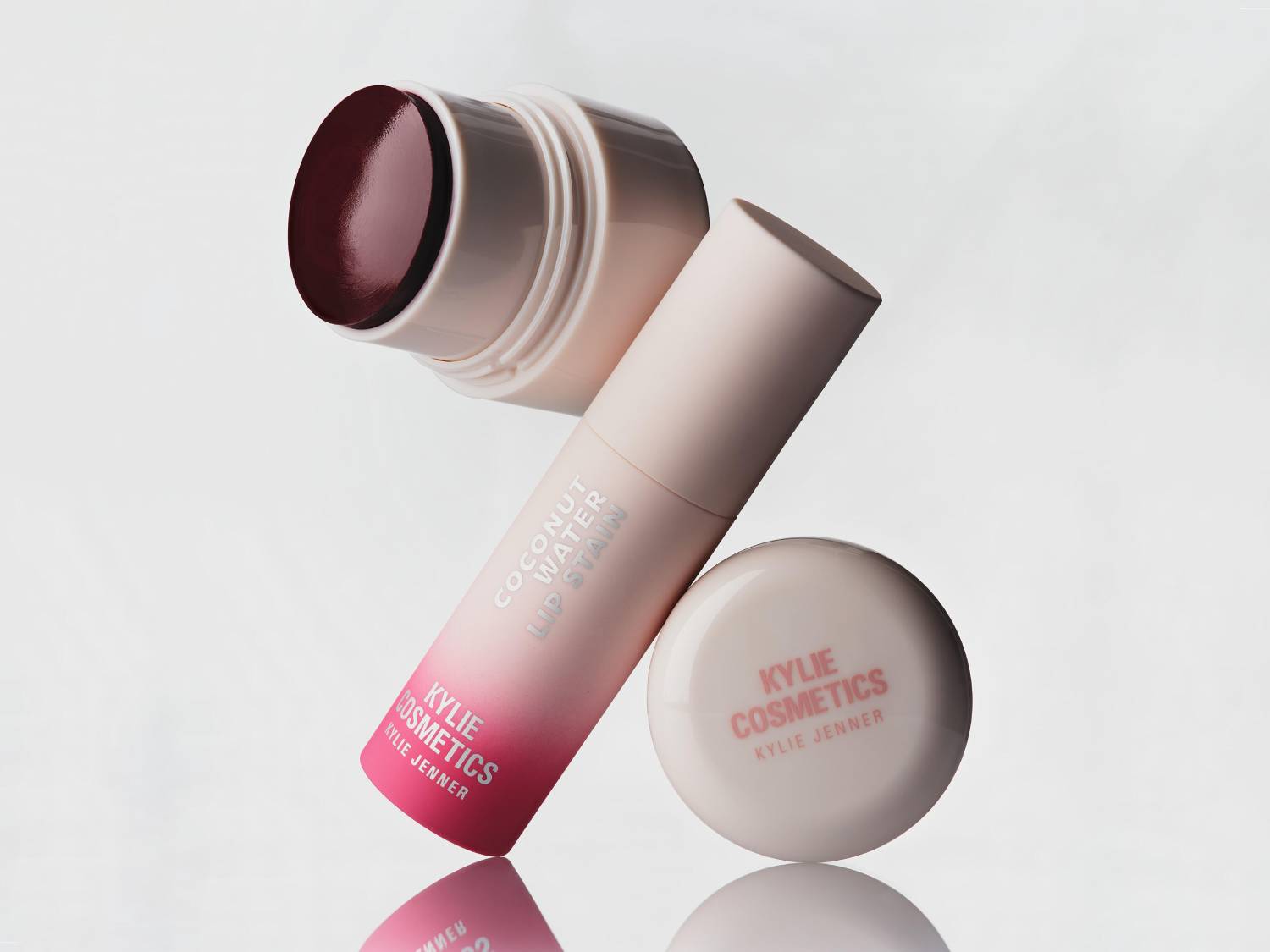 Kylie Cosmetics Coconut Water Lip Stain w odcieniu głębokiego wina, z różowo-białym opakowaniem, leży obok okrągłego słoiczka z logo Kylie Jenner.
