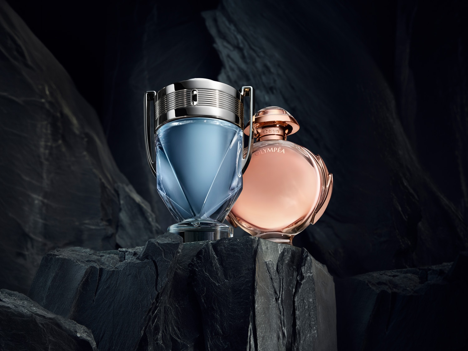 Sticlele de parfum Invictus și Olympea stau pe o stâncă întunecată, Invictus are o sticlă albastră în formă de trofeu, iar Olympea are o sticlă roz-aurie rotundă.
