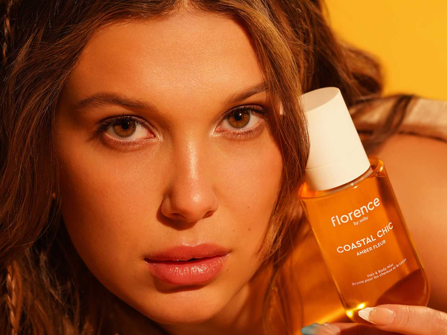 Młoda kobieta prezentuje mgiełkę do włosów i ciała Florence by Mills Coastal Chic Amber Fleur w bursztynowej butelce z białą nakrętką, podkreślając jej promienny wygląd.