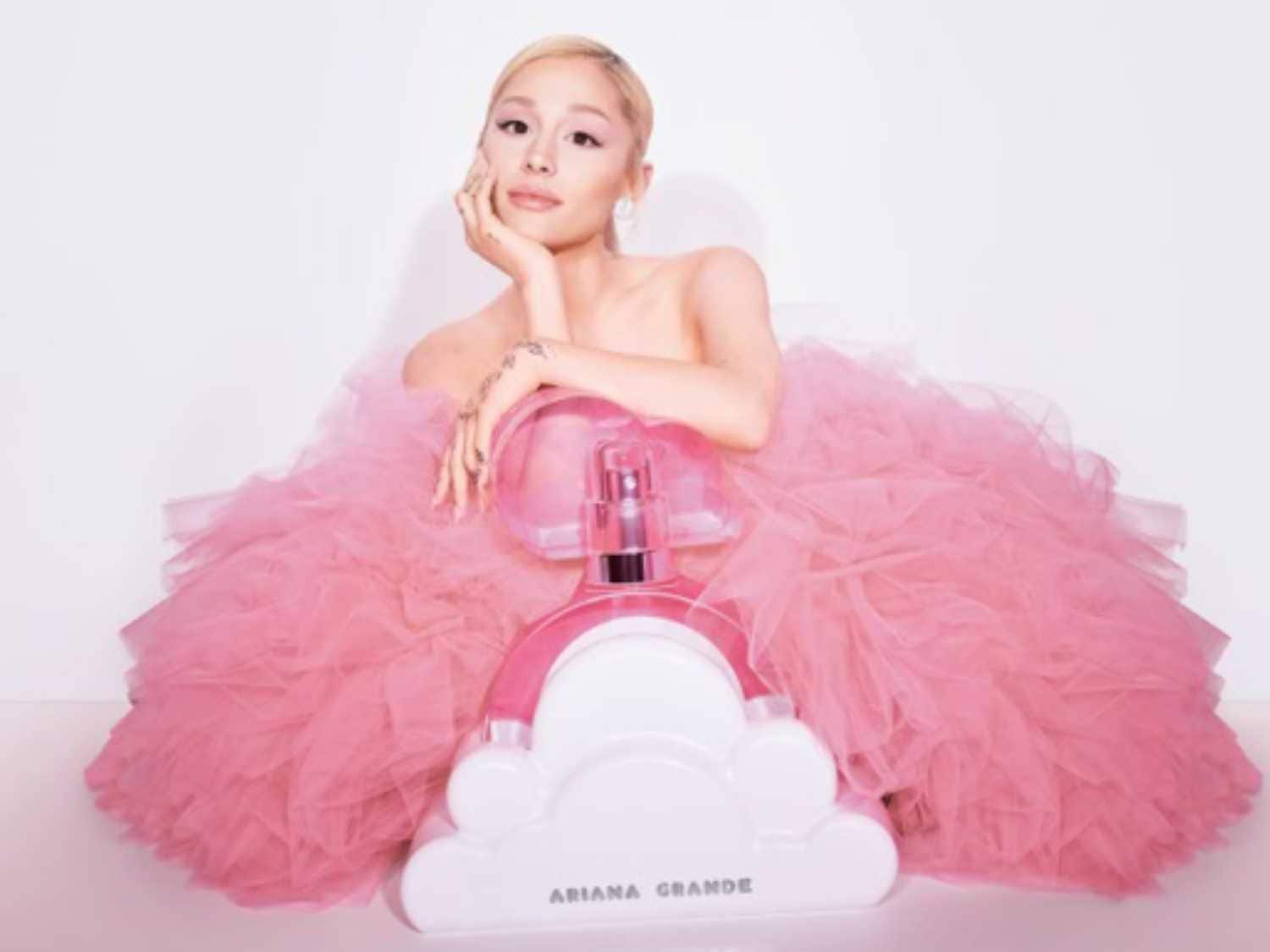 Ariana Grande w różowej sukni z tiulu pozuje obok flakonu perfum w kształcie chmury, tworząc słodki i dziewczęcy wizerunek.