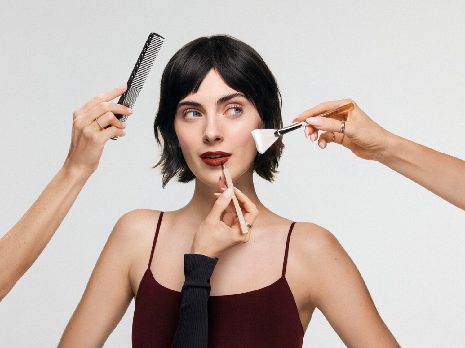 Na portrétu je žena s hnědými vlasy a modrýma očima, líčená rtěnkou a tvářenkou, zatímco jí upravují vlasy hřebenem a nanášejí make-up štětcem.