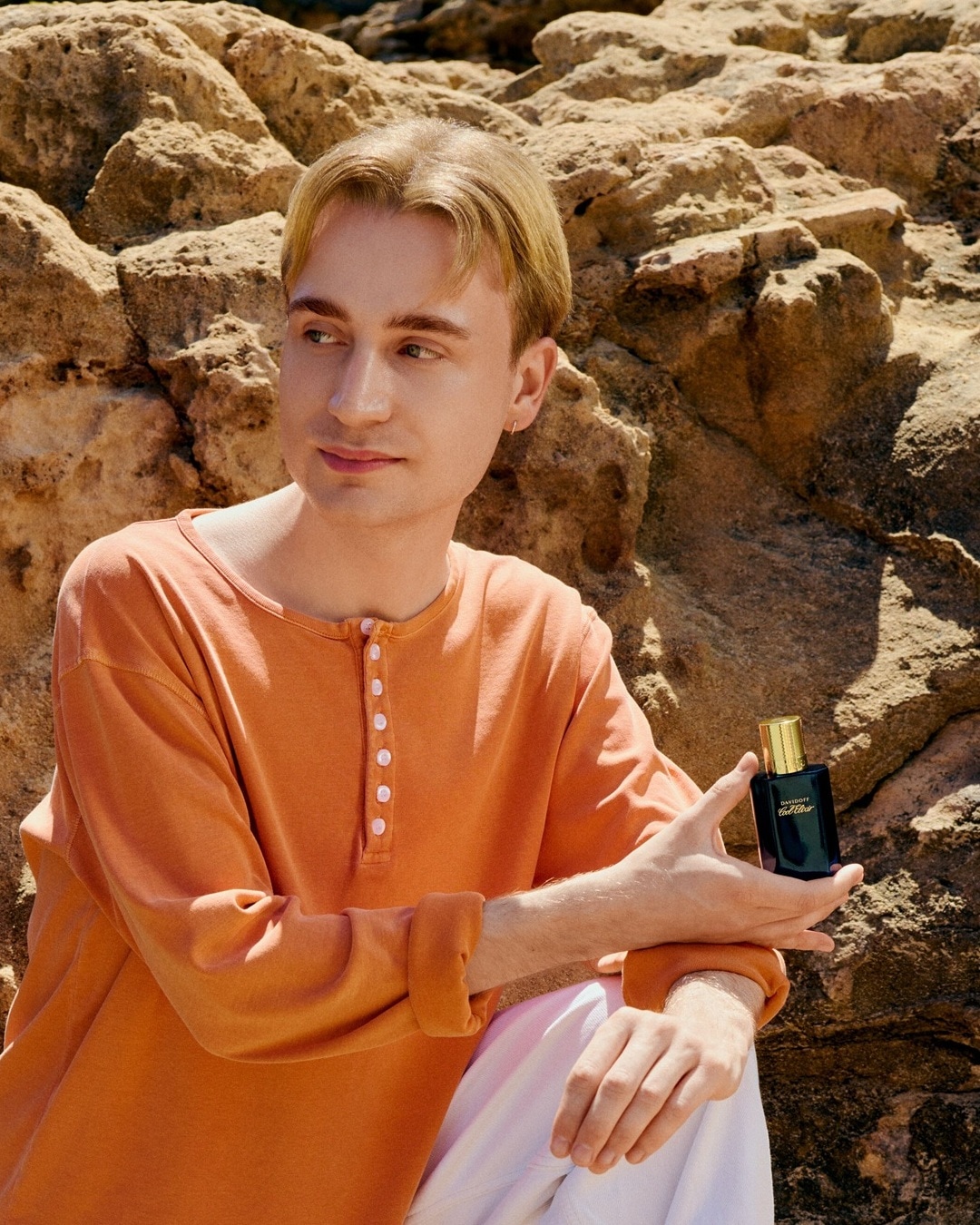 Person in orangefarbenem Oberteil sitzt zwischen Felsen und hält ein dunkles Parfum-Flakon.