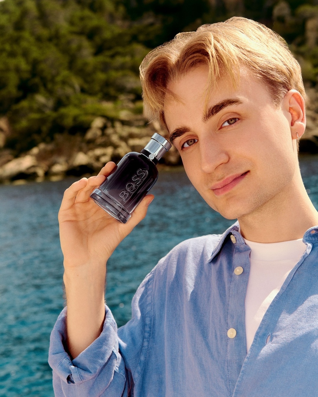 Person hält ein dunkles Parfum-Fläschchen vor dem Meer im Sonnenlicht.