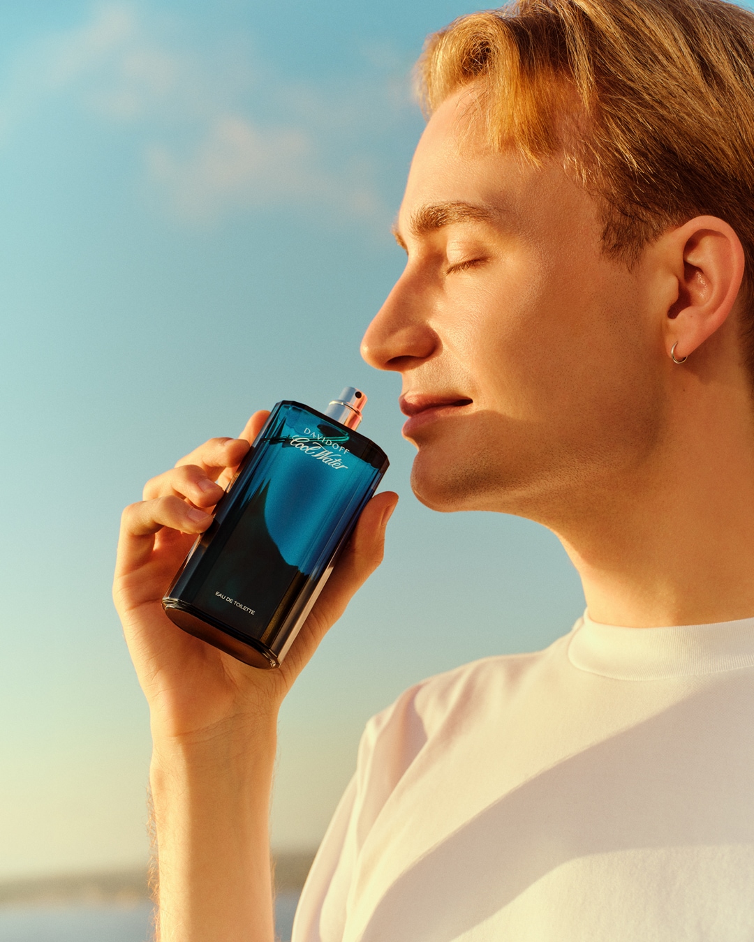 Ein Mann mit blonden Haaren genießt den Duft von Davidoff Cool Water Eau de Toilette, gehalten in einer eleganten, blau getönten Flasche vor einem strahlend blauen Himmel.