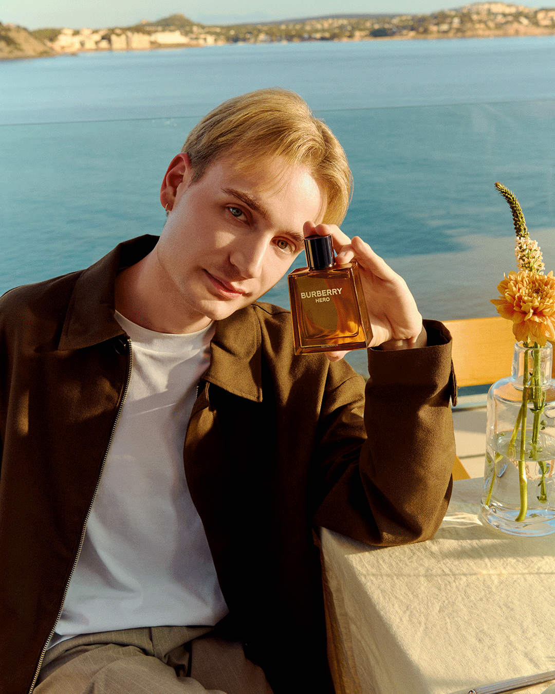 Person sitzt am Tisch am Wasser und hält ein Parfum-Flakon (Burberry) in der Hand.