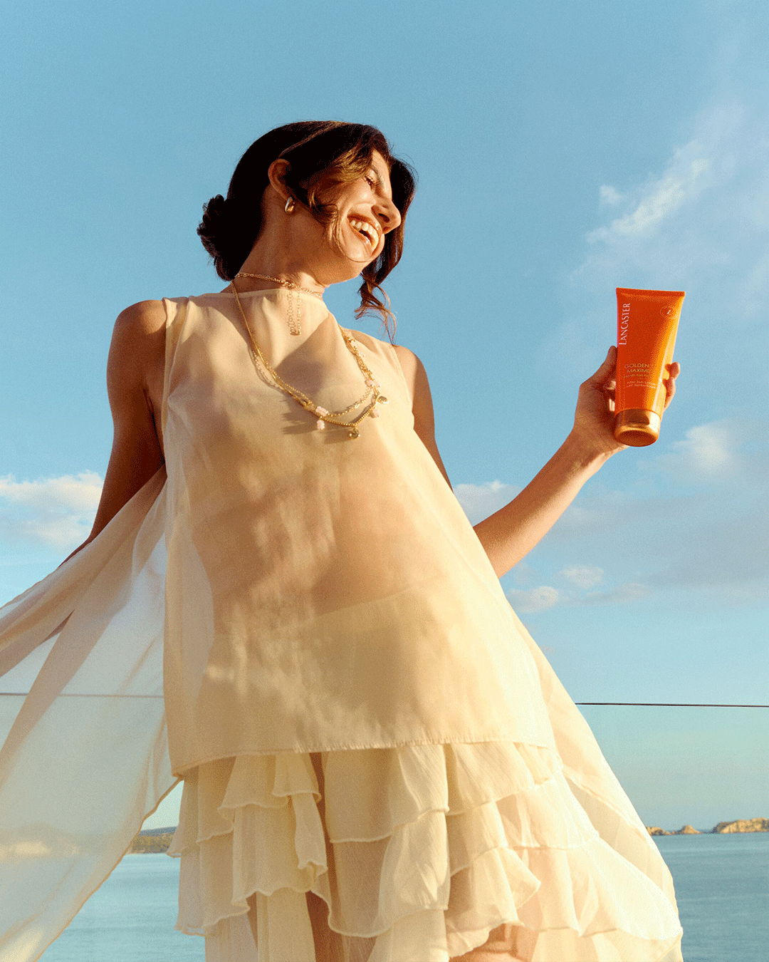 Person in luftigem, hellem Outfit hält eine orangefarbene Sonnencreme vor dem Meer in der Abendsonne.
