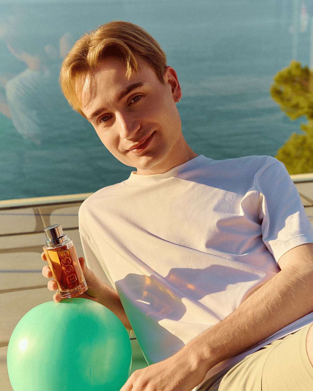 Person sitzt am Meer und hält ein Parfum-Flakon neben einem grünen Ball.