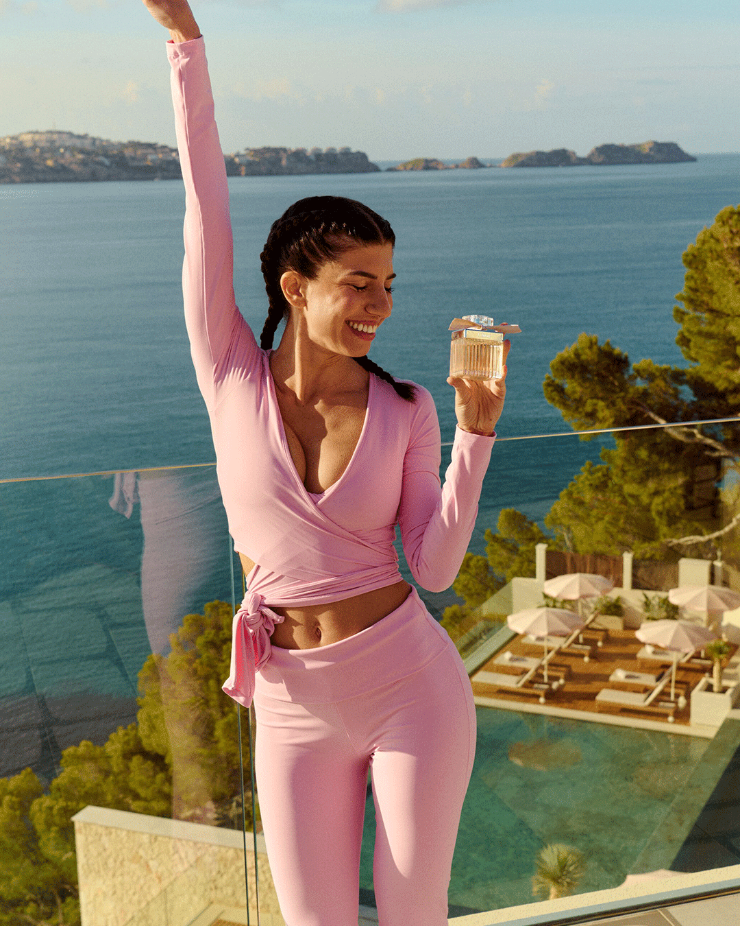 Person im pinken Outfit steht auf einer Terrasse am Meer und hält ein Parfum.