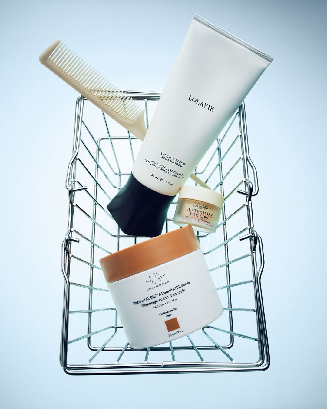 Un panier en fil de fer contient des produits de beauté : un shampoing exfoliant et détoxifiant Lolavie de 180 ml, un masque de nuit pour les lèvres Kiehl's Buttermask et un gommage au lait d'amande Drunk Elephant Sugared Koffie de 270 ml, ainsi qu'un peigne beige.