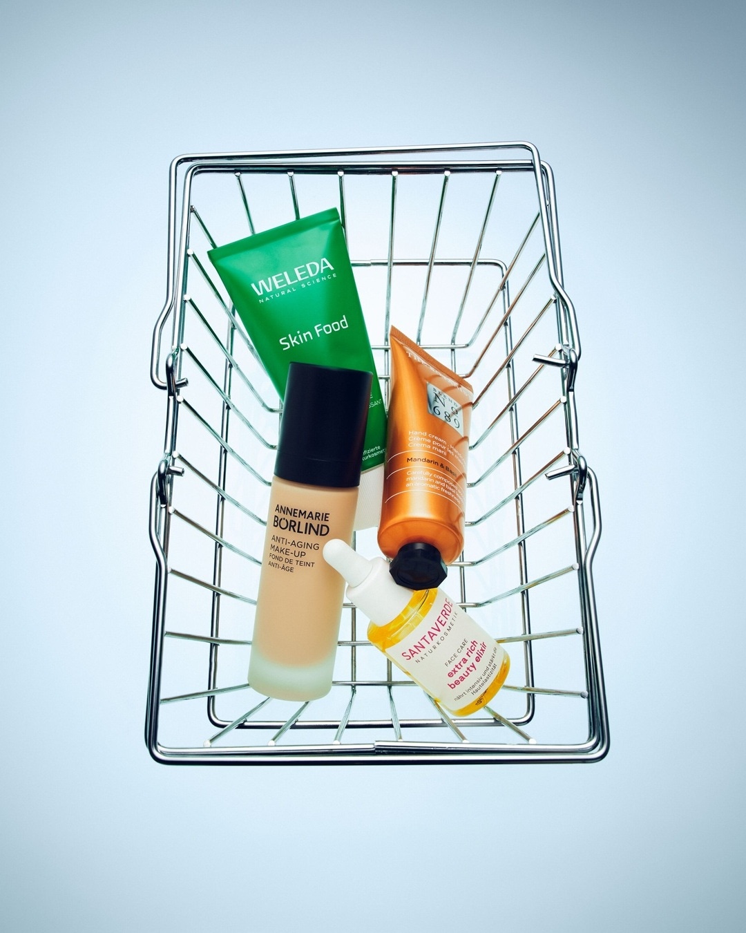 Un panier en fil de fer contient quatre produits de beauté: une crème verte Weleda Skin Food, un fond de teint beige Annemarie Börlind, une crème pour les mains orange Mandarin & Baie et un élixir de beauté jaune Santeverde.