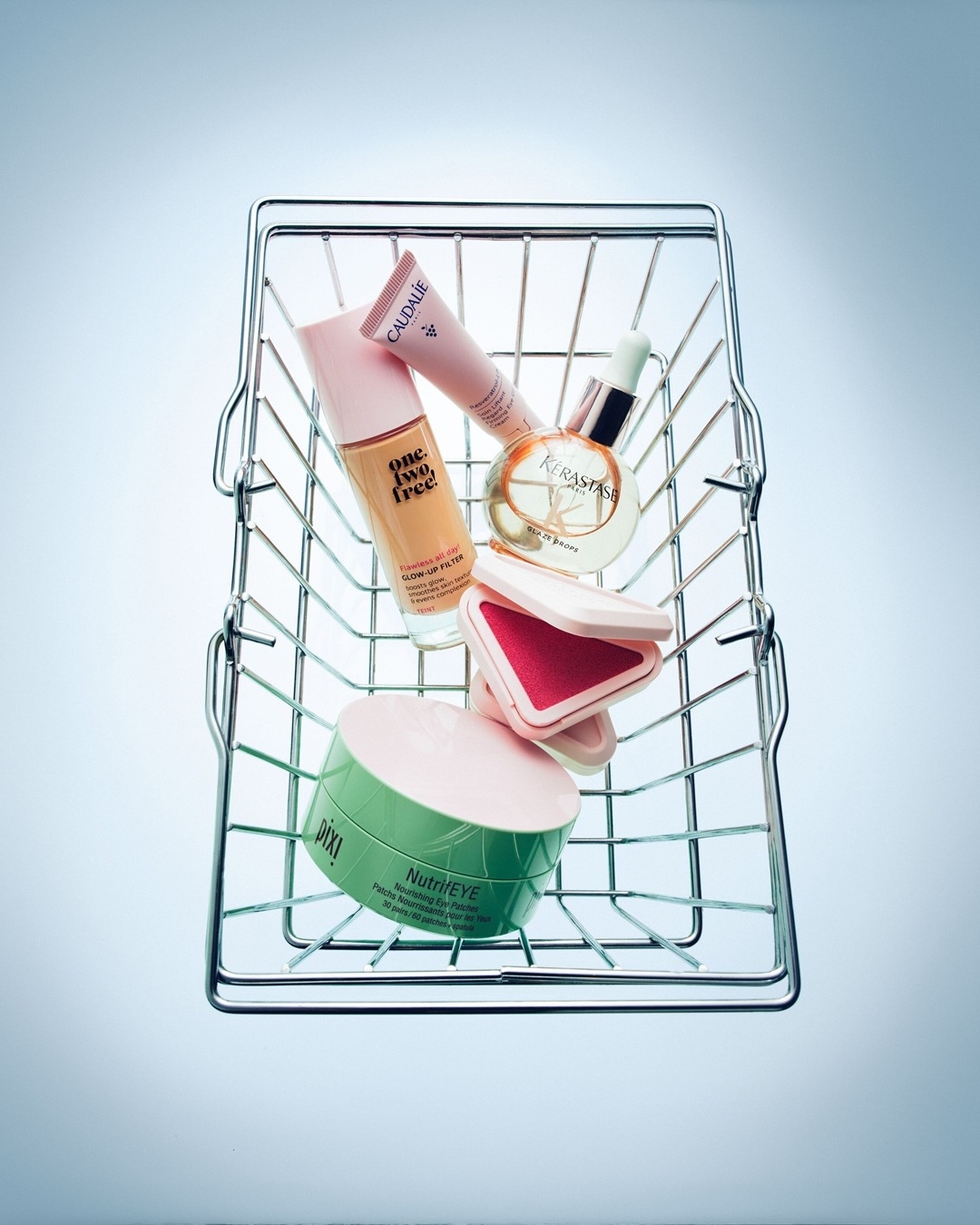 Ein silberner Einkaufskorb gefüllt mit Schönheitsprodukten wie One. Two. Free! Glow-Up Filter, Caudalie Resveratrol Lift Augenlifting Creme, Kérastase Glaze Drops und Pixi NutrifEYE Augenpads.