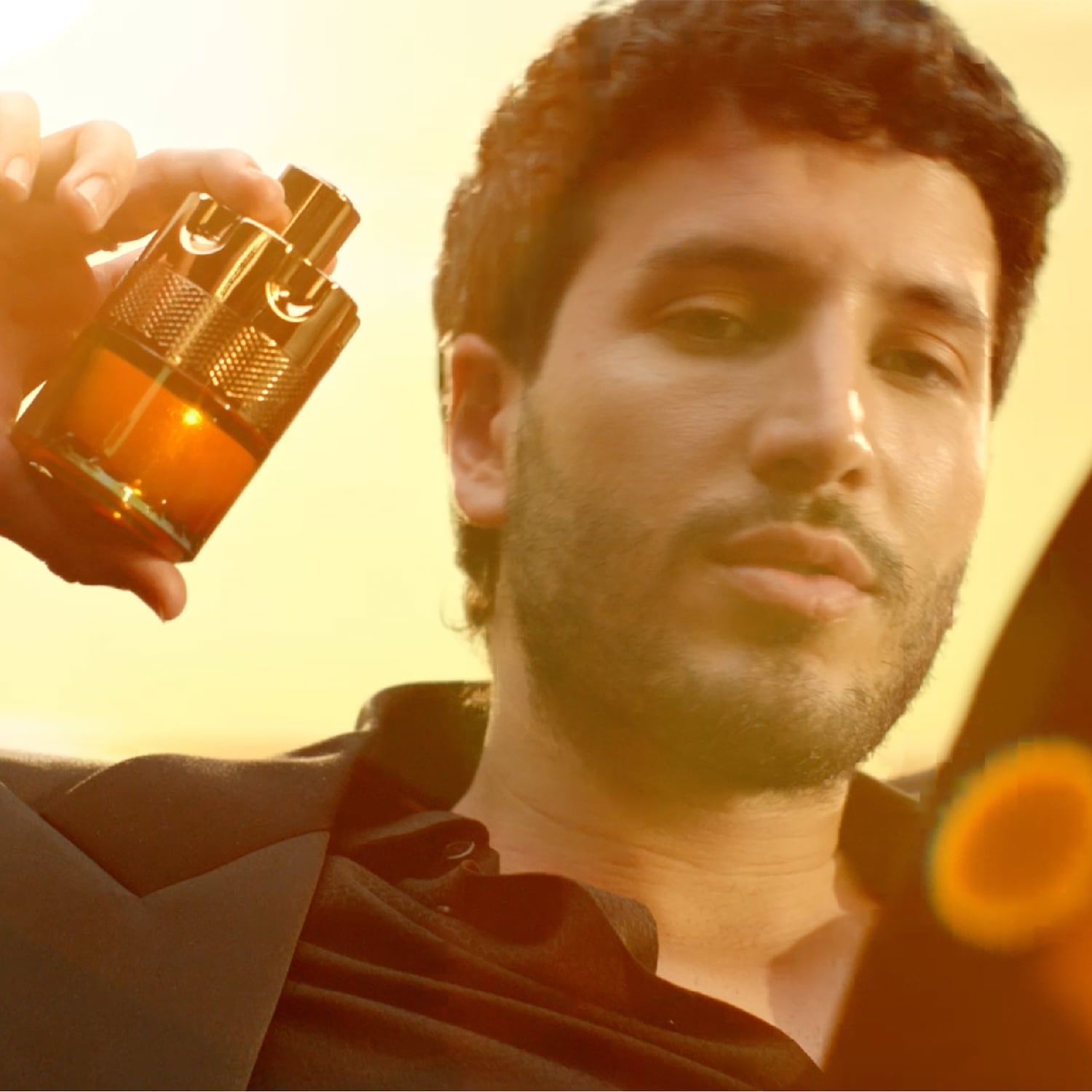 Un homme tient un flacon de parfum ambré et doré, le soleil couchant créant une ambiance chaleureuse et luxueuse.