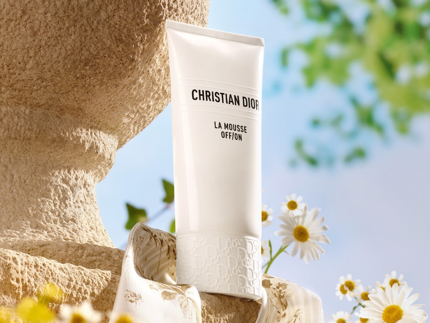 De Christian Dior La Mousse OFF/ON gezichtsreiniger in een witte tube met zwarte letters staat op een beige stenen sokkel, omringd door witte madeliefjes en groene bladeren tegen een helderblauwe lucht.