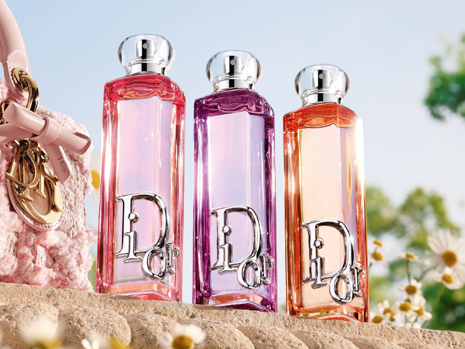 Drie langwerpige Dior-parfumflesjes in roze, paars en oranje staan op een zandkleurige ondergrond met bloemen, met een roze tasje ernaast.