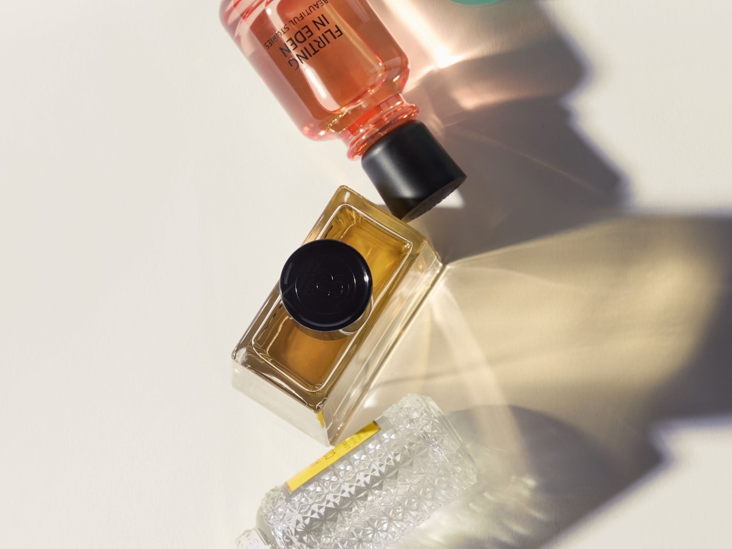 Drei Parfümflaschen liegen auf einer hellen Oberfläche, darunter ein rechteckiger Flakon mit goldener Flüssigkeit und schwarzem Deckel sowie eine Flasche mit rosa Flüssigkeit.
