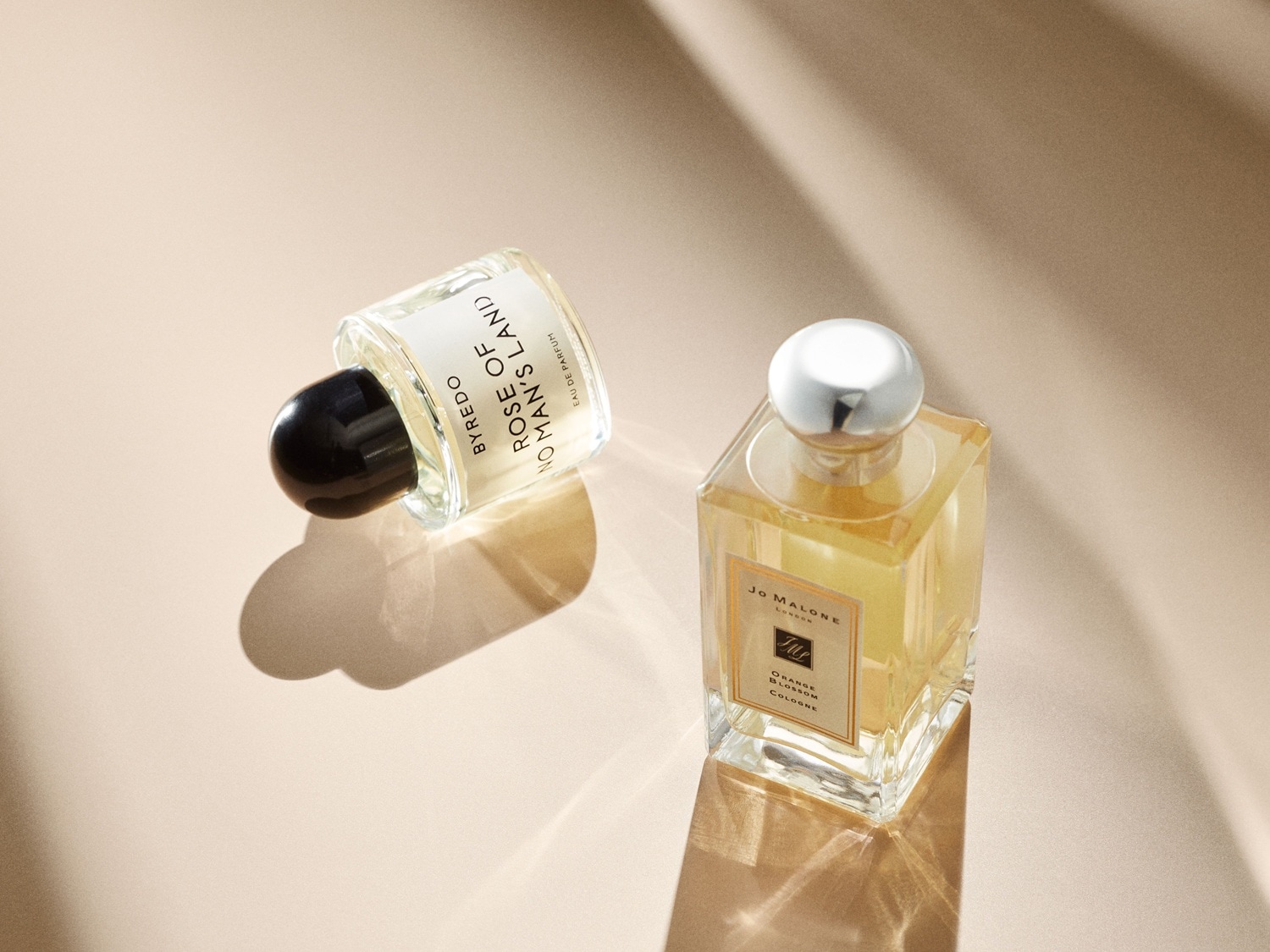 Zwei Parfümflaschen liegen auf einer hellbraunen Oberfläche, eine von Byredo Rose of No Man's Land mit schwarzem Verschluss und eine von Jo Malone Orange Blossom mit silbernem Verschluss.