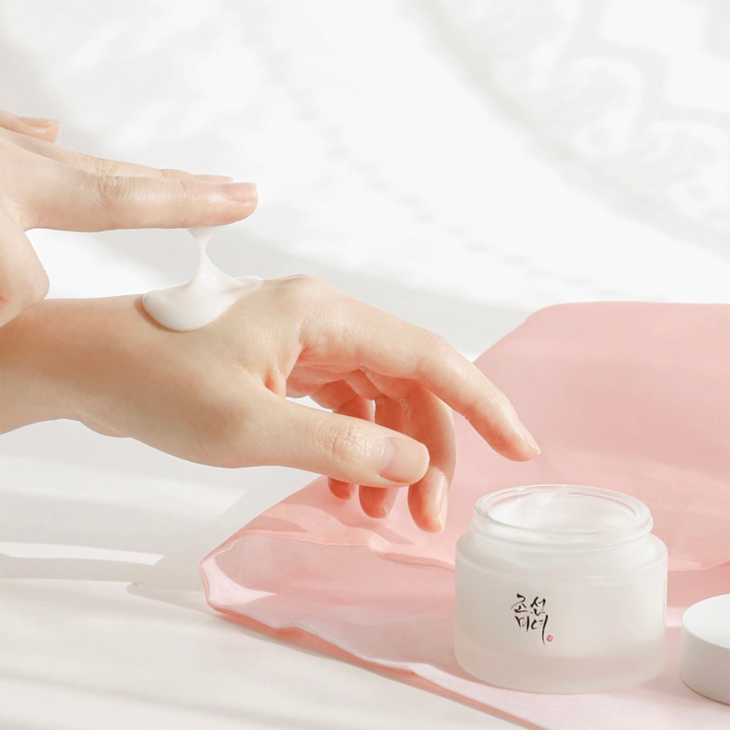 Una mano applica una crema bianca e densa da un barattolo di vetro trasparente su un panno rosa chiaro, mostrando la consistenza idratante del prodotto.