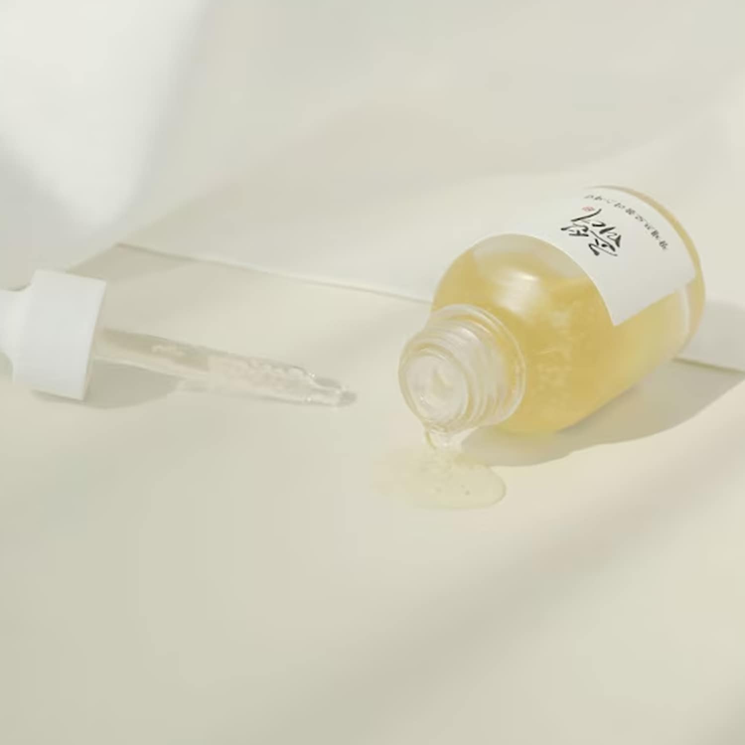 Flacone di siero dorato disteso su un fianco con contagocce bianco e liquido che fuoriesce, per una pelle luminosa e idratata.