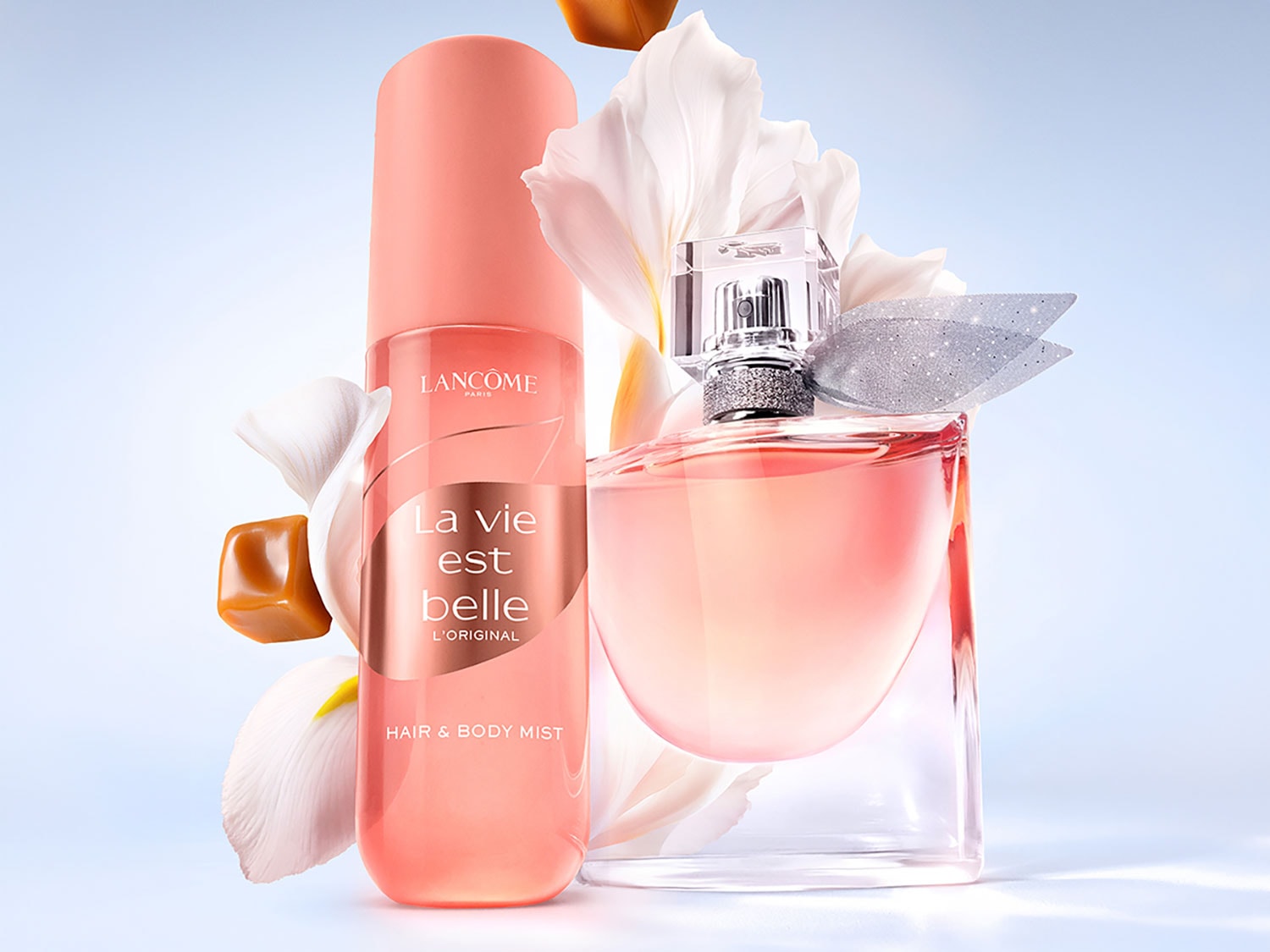 Spray-ul parfumat pentru păr și corp La Vie Est Belle L'Original de la Lancôme, într-un recipient roz piersică, alături de parfumul cu același nume, flori albe și caramelă.