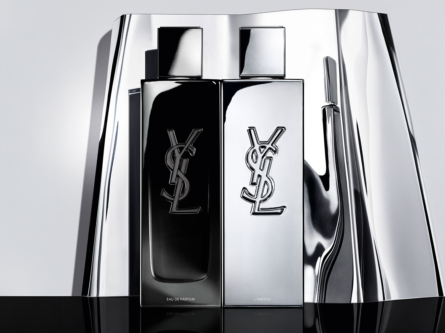 Două sticle de parfum YSL, una neagră și una argintie, cu logo-ul YSL proeminent, prezentate pe un fundal metalic lucios, emanând eleganță și rafinament.