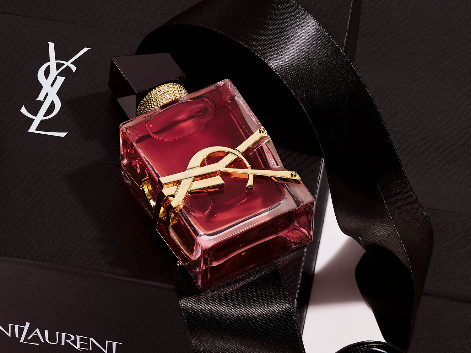 Sticlă de parfum YSL Libre roșu-rubiniu, cu logo-ul auriu YSL, așezată pe o pungă neagră de cumpărături, accentuată cu o panglică neagră lucioasă, simbolizând lux și eleganță.